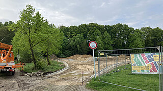 Baustelle mit unbefestigtem Weg, Bauzaun mit bunter Werbetafel und Verkehrsschild vor einem Wald