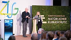Ein Mann und eine Frau stehen auf einer Bühne, rechts von ihnen eine Mooswand mit der Aufschrift "Kompetenzzentrum Natürlicher Klimaschutz"