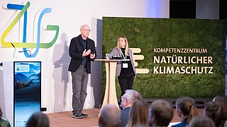 Ein Mann und eine Frau stehen auf einer Bühne, rechts von ihnen eine Mooswand mit der Aufschrift "Kompetenzzentrum Natürlicher Klimaschutz"