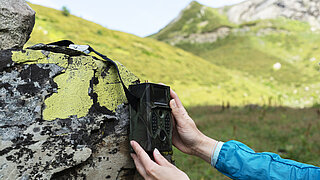 Das Bild zeigt zwei Hände, die eine Kamera oder Messgerät an einen Felsblock halten, der mit Flechten bedeckt ist, im Hintergrund eine Berglandschaft.