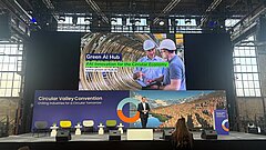 Ein Mann steht auf einer Bühne, im Hintergrund sieht man mehrere Bildschirme