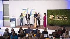 Fünf Personen stehen auf einer Bühne vor Publikum, drei Männer und zwei Frauen, neben einem grünen Mooswand-Logo mit der Aufschrift 'Kompetenzzentrum Natürlicher Klimaschutz'.