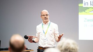 Ein Mann spricht zu einem Publikum. Im Hintergrund sieht man eine Powerpoint-Präsentation