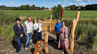 Fünf Personen stehen auf einer Holzplattform neben geschnitzten Holzfiguren in einer Wiese unter blauem Himmel