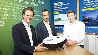 Drei Männer stehen um einen Staubsaugroboter mit Digitalem Produktpass, der direkt mit einem KI-Chatbot kommuniziert. 