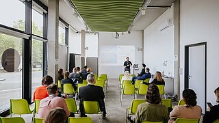 Mehrere Personen sitzen auf Stühlen. Vor ihnen steht eine Person und spricht. 