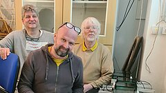 3 Männer knien neben Geräten. Einer hält ein Laptop 