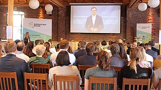 Ein Mann (BM Carsten Schneider) ist auf einer Leinwand zu sehen, vor ihm sitzt ein Publikum