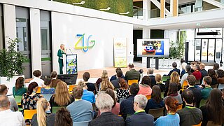 Eine Person steht auf der Bühne und spricht, vor ihr sitzt ein Publikum auf Stühlen. 