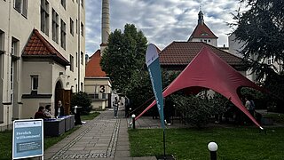 Weg mit Infostand und blauem Willkommensschild links sowie rotem Zelt und Fahne rechts vor Gebäuden und Bäumen