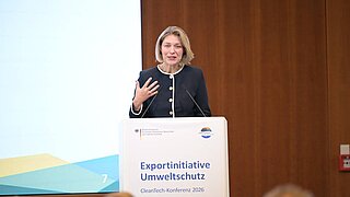 Eine Frau steht hinter einem Rednerpult und spricht in ein Mikro. 