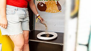 Ene Frau zeigt auf eine Trockentoilette, die ohne Wasserspülung funktioniert.