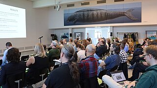 Mehrere Personen sitzen auf Stühlen in einem Raum und schauen auf eine Präsentation, die an der Wand sichtbar ist. 