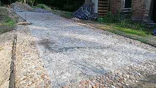 Weg mit gepflasterter Fläche aus rechteckigen Steinen und Rand aus runden Steinen vor einem Backsteingebäude