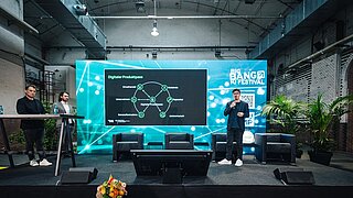Drei Männer auf einer Bühne vor großem Bildschirm mit digitalem Produktpass und Logo des Big Bang KI Festivals in einer Industriehalle