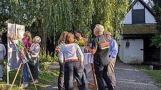 Mehrere Personen stehen an einem Stehtisch, daneben schauen sich zwei weitere Personen eine Infotafel an. 