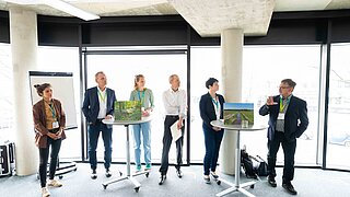 Mehrere Personen befinden sich an Stehtischen und sprechen miteinander. 