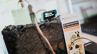 ein Sensor steckt in einem Topf voll Pflanzenkohle, im Vordergrund ein Flyer der KI-Ideenwerkstatt