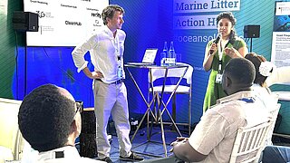 Zweo Personen stehen und sprechen zu einem Publikum, das vor ihnen sitzt