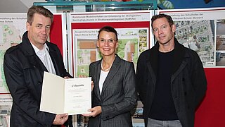 Drei Personen stehen vor einer aufklappbaren roten Pinnwand, die Personen links und in der Mitte halten eine Urkunde