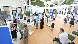 Mehrere Personen befinden sich im Foyer eines Gebäudes, sprechen miteinander und sehen sich Anschauungsmaterialien (Poster etc.) an. 