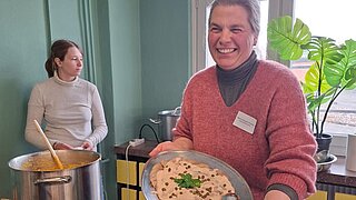 Zwei Personen stehen vor einem Tisch, eine hält einen Teller mit etwas zu Essen in den Händen. Auf dem Tisch befinden sich ein Kochtopf und weitere Teller.