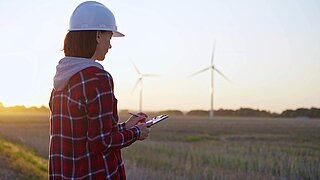 Person mit Schutzhelm und Klemmbrett steht vor offenem Feld mit Windkraftanlagen