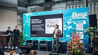 Drei Männer auf Bühne vor großem Bildschirm mit Präsentationstext und QR-Code beim Big Bang Festival in Industriehalle.