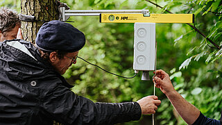 Drei Personen prüfen den Abstand von einem Gerät zum darunterliegenden (nicht sichtbaren) Bach. 