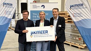 Drei Personen halten gemeinsam ein Schild mit der Aufschrift "BK Meer" in den HÄnden
