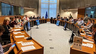 Viele Personen sitzen in einem Konferenzraum um einen U-förmigen Tisch herum, vor ihnen liegen Unterlagen 
