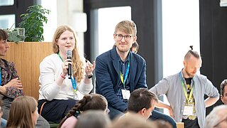 Drei Personen sitzen/stehen im Publikum. Eine hält ein Mikrofon und spricht. 