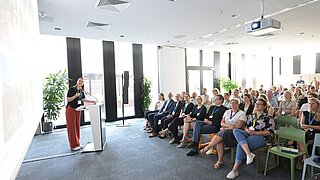 Eine Person steht an einem Rednerpult und spricht. Vor ihr Stuhlreihen mit Publikum