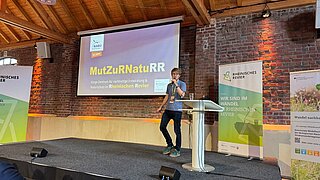 Eine Person steht auf einer Bühne, auf der Bühne sind eine Projektionsfläche, Rollups und ein Rednerpult