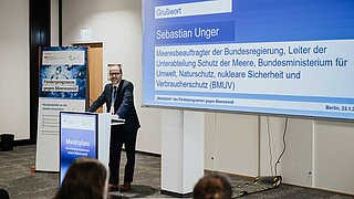 Ein Mann steht an einem Rednerpult, hinter ihm befindet sich ein großer Bildschirm, auf dem eine Präsentation zu sehen ist. 