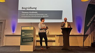Zwei Personen auf Bühne vor Leinwand mit Text 'Begrüßung Thomas Kralinski, Leiter der Abteilung Nationaler und europäischer Klimaschutz im BMUKN'