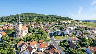 Blick auf der Luft auf ein Dorf mit Häusern und Straßen, im Hintergrund bewaldete Hügel 