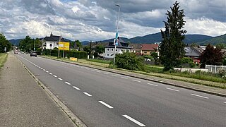 eine Straße durch eine Ortschaft, an einer Seite ein Gehweg, auf der anderen Seite Häuser und einzelne Bäume und Büsche 