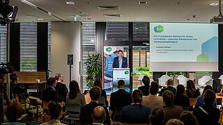 Blick in den gefüllten Konferenzraum zum KEI Podium, auf der Bühne spricht Benjamin Görlach.
